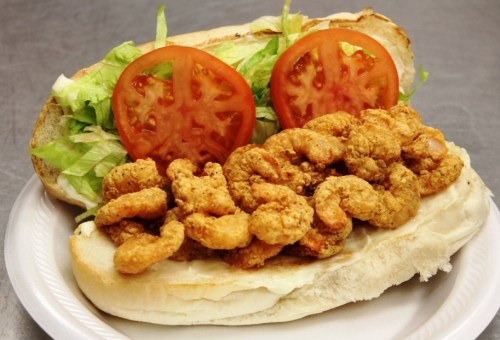 A sandwich filled with fried shrimp, lettuce, and tomato slices on a hoagie roll, served on a white plate.