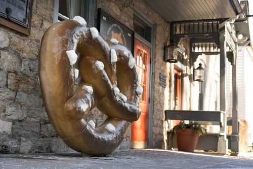 A large, decorative pretzel sculpture stands outside a stone building, with a potted plant nearby.