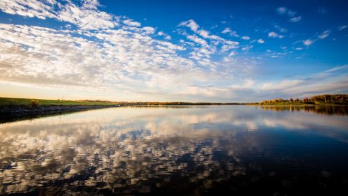 A serene lake reflecting a blue sky with clouds, surrounded by green fields and trees.
