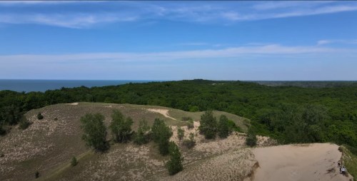 Aerial view of lush green hills leading to a sandy area, with a glimpse of a blue lake in the background under a clear sky.