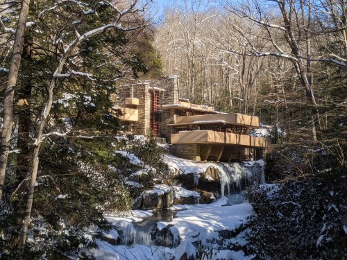 A modern house with multiple levels, nestled among snowy trees and a frozen waterfall in a serene winter landscape.