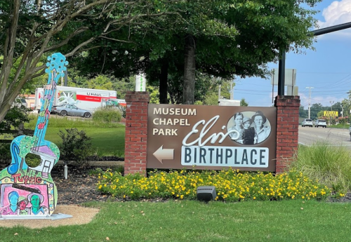 Sign for Elvis Presley’s birthplace, featuring colorful guitar art and surrounded by greenery and flowers.