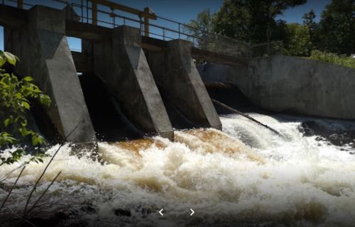 A concrete dam with water flowing rapidly over it, surrounded by greenery and trees.