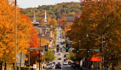 A vibrant autumn scene of a tree-lined street with colorful foliage, shops, and traffic lights in a small town.