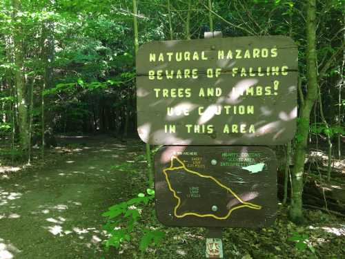 A wooden sign in a forest warns of falling trees and limbs, advising caution in the area. A trail map is also visible.