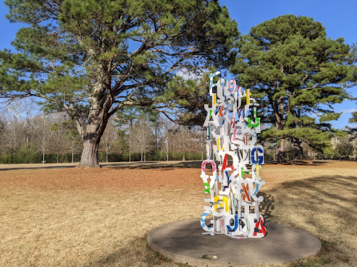 Colorful alphabet letters stacked vertically in a park, surrounded by tall trees and a grassy area.