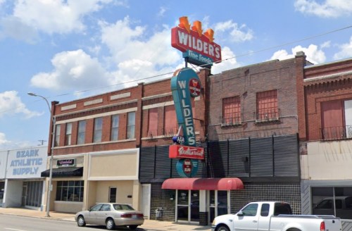A vintage sign for Wilders stands tall above a brick building, with a clear blue sky and a few cars parked nearby.