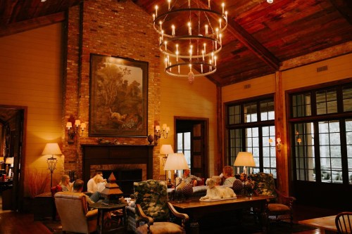 Cozy living room with a large chandelier, stone fireplace, and people gathered around, with a dog resting nearby.