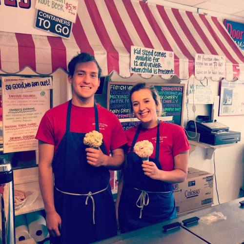 Two smiling employees hold ice cream cones in a colorful shop with a striped awning and various signs in the background.
