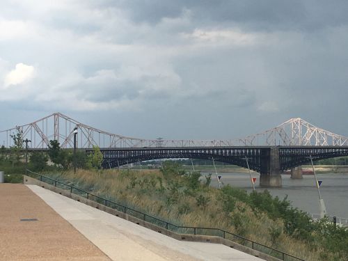 A bridge spans a river under a cloudy sky, with greenery and a pathway in the foreground.
