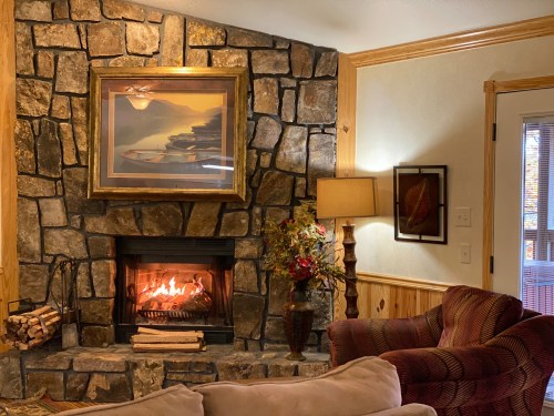 Cozy living room with a stone fireplace, framed artwork, a lamp, and a comfortable sofa. Warm ambiance with a fire.