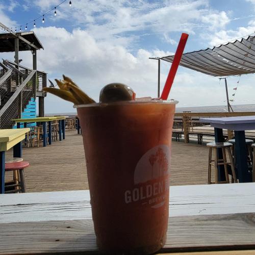 A cocktail with a straw and garnish sits on a table, with a scenic view of the ocean and cloudy sky in the background.