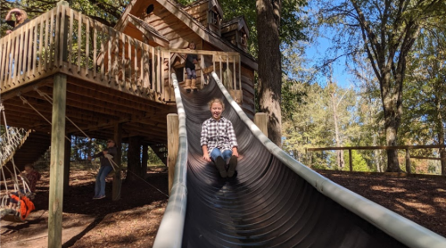 A child slides down a large slide from a wooden treehouse, surrounded by trees and other children playing.