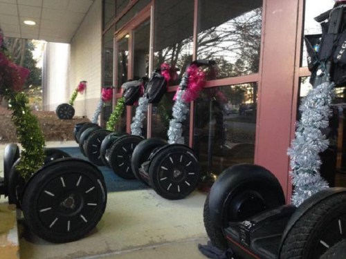 A row of decorated Segways with festive garlands and pink bows outside a building entrance.