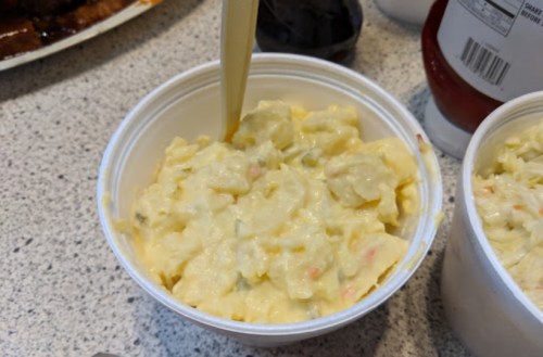 A close-up of a container filled with creamy potato salad, with a spoon resting inside.