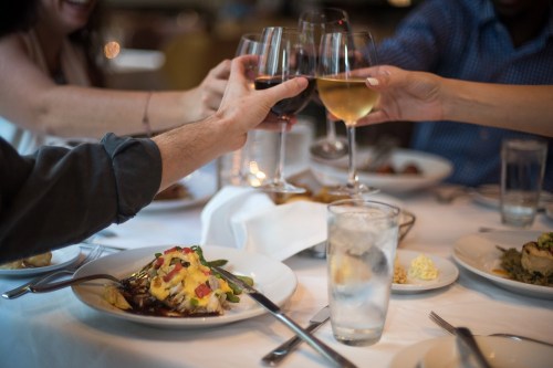 A group toasts with wine glasses over a dining table with plates of food and water glasses.