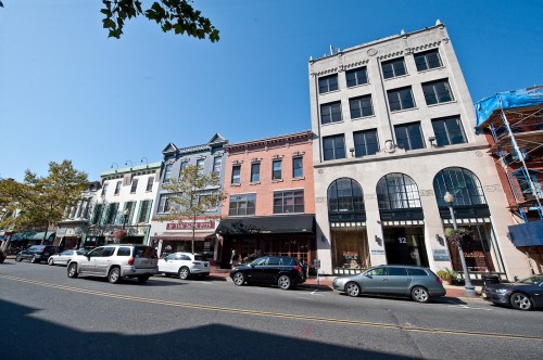 A street view featuring historic buildings, shops, and parked cars under a clear blue sky.