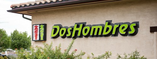 Sign for "Dos Hombres" with colorful graphics, mounted on a light-colored building, surrounded by greenery.