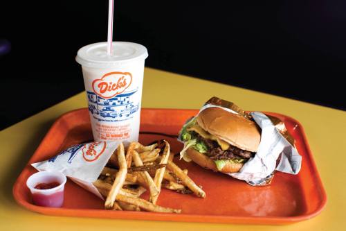 A tray with a burger wrapped in foil, fries, a cup of soda, and a small container of ketchup on a yellow table.