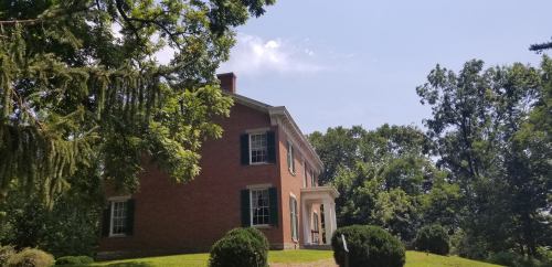 A historic brick house with green shutters, surrounded by trees and a well-maintained lawn under a clear blue sky.