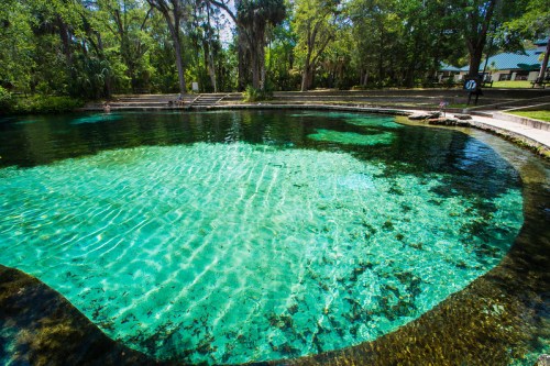 A serene, clear blue spring surrounded by lush greenery and stone pathways, inviting for swimming and relaxation.