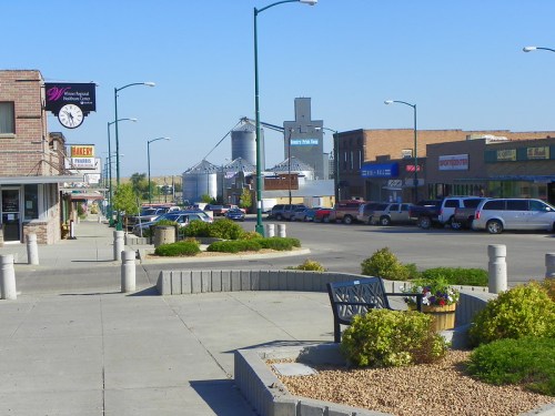 A small town street lined with shops, parked cars, and greenery under a clear blue sky.