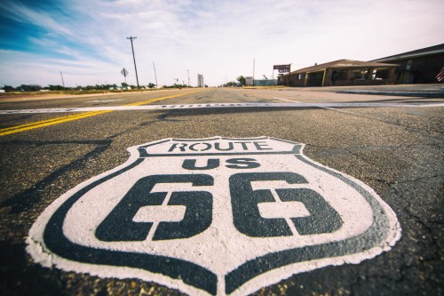 A close-up of a Route 66 sign painted on the road, with a clear blue sky and distant buildings in the background.