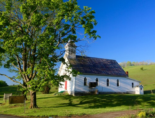 A quaint white church with a steeple, surrounded by green grass and a large tree, under a clear blue sky.