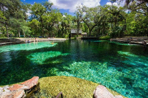 A serene natural spring with clear turquoise water, surrounded by lush greenery and people enjoying the scenery.