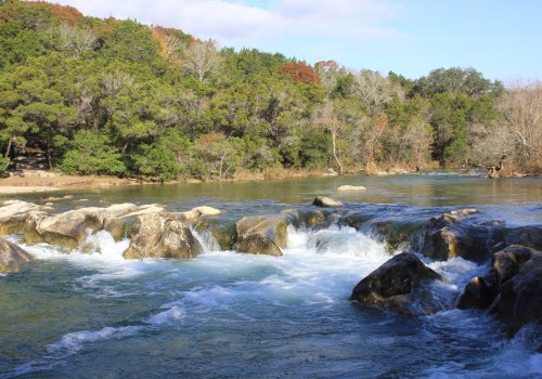 A serene river flows over rocks, surrounded by lush green trees and a clear blue sky.