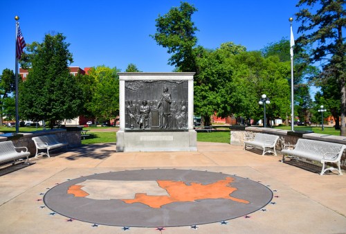A memorial with a bronze relief depicting historical figures, surrounded by benches and trees in a park setting.