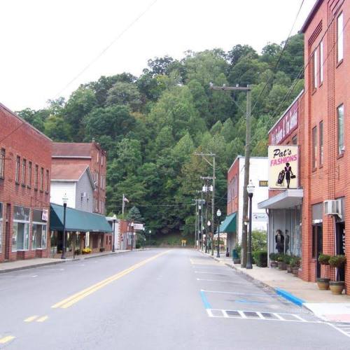 A quiet street lined with brick buildings and shops, set against a backdrop of green hills.