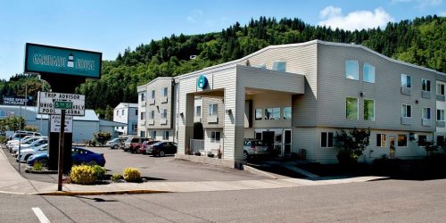 Exterior view of a hotel with a parking lot, surrounded by greenery and a clear blue sky.