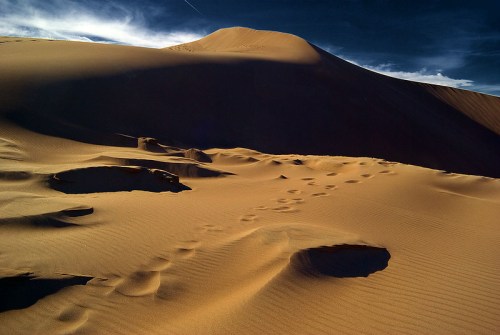 A vast desert landscape with gentle sand dunes and scattered footprints under a dramatic sky.