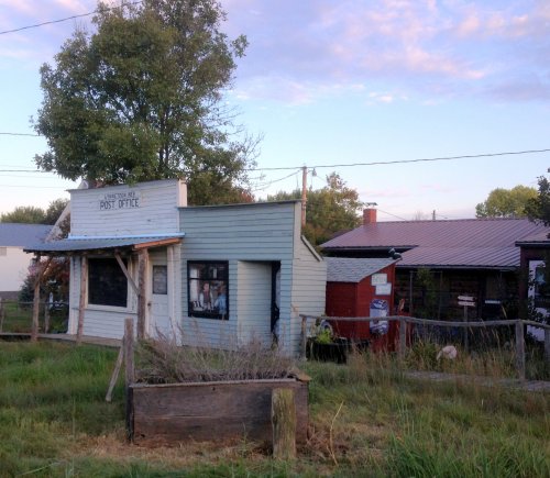 A small, rustic post office building surrounded by grass and trees, with a red building nearby under a colorful sky.
