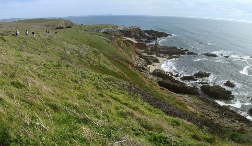 A coastal landscape with grassy hills, rocky shores, and a calm ocean under a clear sky, with people walking along the path.