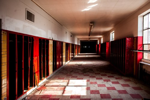 Abandoned school hallway with red and yellow lockers, sunlight streaming through windows, and a checkered floor.