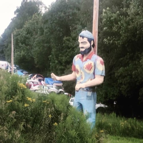 A large, colorful statue of a man in work clothes stands beside a road, surrounded by greenery and parked cars in the background.