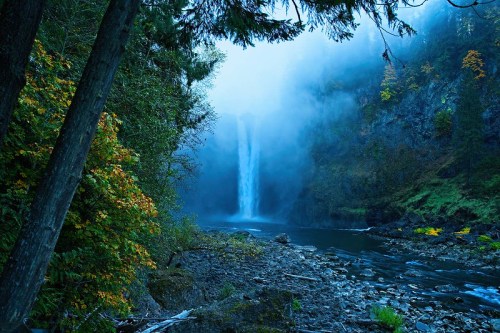 A misty waterfall cascades down rocky cliffs, surrounded by lush greenery and autumn-colored trees.