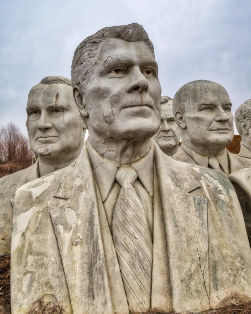 A group of weathered, oversized busts of men in suits, partially eroded, against a cloudy sky.