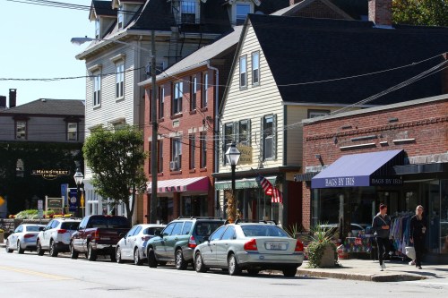 A charming street lined with shops and parked cars, featuring a mix of brick and wooden buildings under clear blue skies.