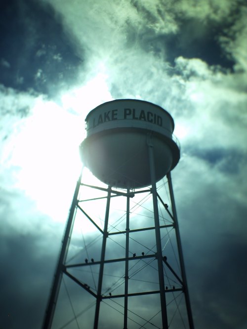 A silhouette of a water tower labeled "Lake Placid" against a cloudy sky with sunlight peeking through.