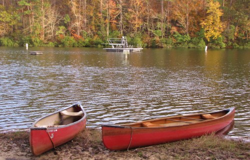Two red canoes on a sandy shore, with a calm lake and a boat in the background surrounded by autumn foliage.