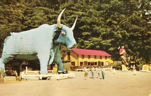 A giant blue bull statue stands beside a yellow building, with people and trees in the background.