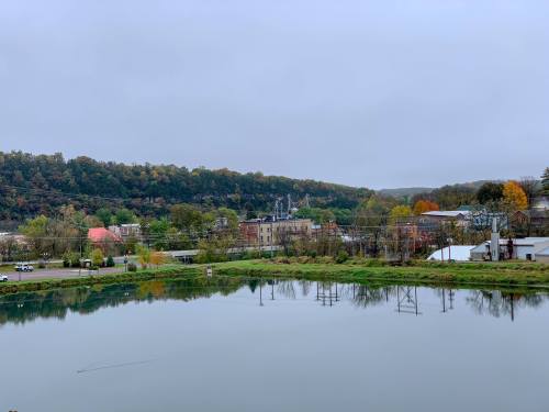A calm river reflects a small town surrounded by green hills under a cloudy sky.
