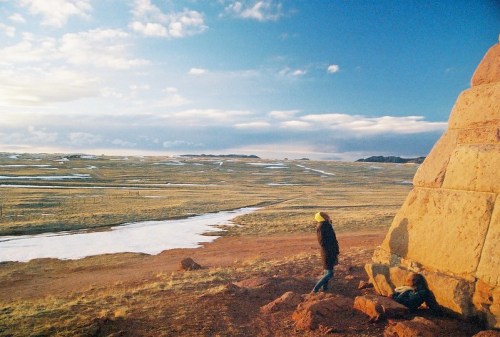 Two people stand near a large rock formation, overlooking a vast, open landscape with patches of snow and a blue sky.