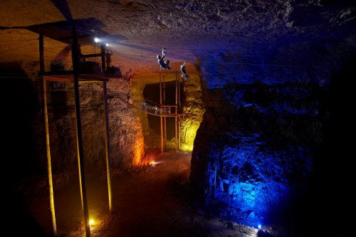 Two people ziplining in a dimly lit cavern with colorful lights illuminating the rock formations.
