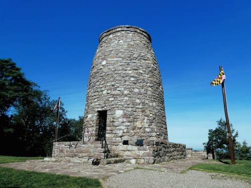 Stone tower on a grassy hill, surrounded by trees, under a clear blue sky, with flags nearby.