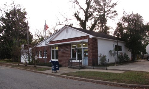 A small brick building with a white facade, featuring a flag and a mailbox outside, set in a quiet street.