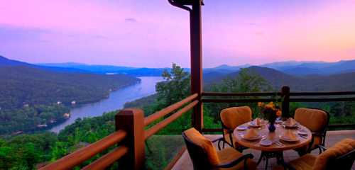 A scenic view from a balcony overlooking a lake and mountains at sunset, with a dining table set for a meal.